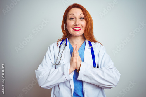Young redhead doctor woman using stethoscope over white isolated background praying with hands together asking for forgiveness smiling confident.