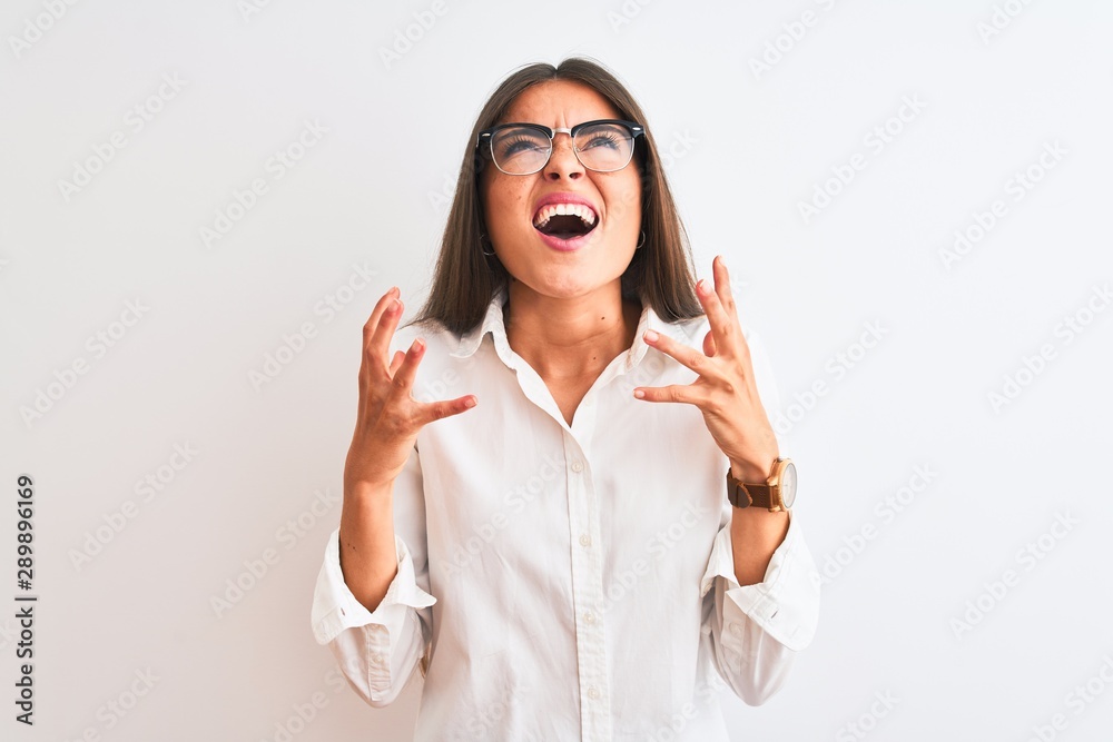Young beautiful businesswoman wearing glasses standing over isolated white background crazy and mad shouting and yelling with aggressive expression and arms raised. Frustration concept. Young beautiful businesswoman wearing glasses standing over isolated white background crazy and mad shouting and yelling with aggressive expression and arms raised. Frustration concept.