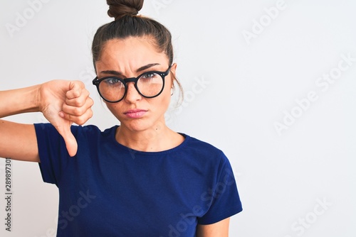 Beautiful woman with bun wearing blue t-shirt and glasses over isolated white background with angry face  negative sign showing dislike with thumbs down  rejection concept