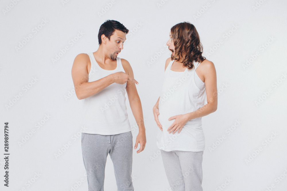 pregnant woman and man in sportswear on white background. The girl holds her stomach with her mouth wide open and a surprised on her face