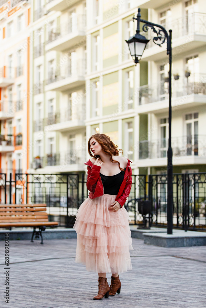 Young curled girl weared in tulle skirt and red leather jacket on background of the City near old classic lantern. Fashion style girl with long curled red hair.  Street style Modern strict urban girl