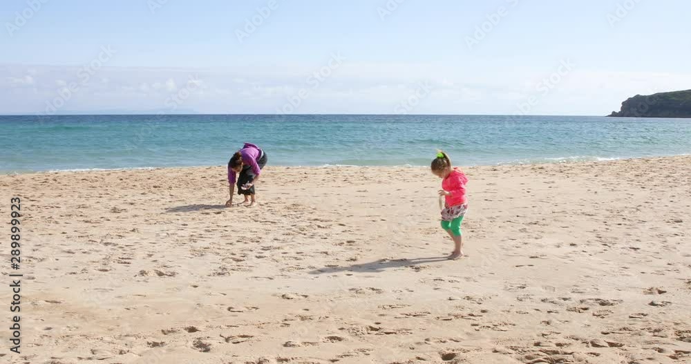 four years old girl picking bone of squid and collecting sea shells at sand beach shore, next to mother or babysitter, in Bolonia Beach, Cadiz, Andalusia, Spain, Europe. four years old girl picking bone of squid and collecting sea shells at sand beach shore, next to mother or babysitter, in Bolonia Beach, Cadiz, Andalusia, Spain, Europe.