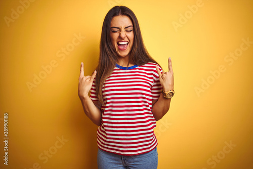 Young beautiful woman wearing striped t-shirt standing over isolated yellow background shouting with crazy expression doing rock symbol with hands up. Music star. Heavy concept.