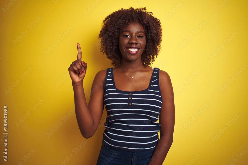 Young african afro woman wearing striped t-shirt over isolated yellow background showing and pointing up with finger number one while smiling confident and happy.