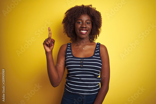 Young african afro woman wearing striped t-shirt over isolated yellow background showing and pointing up with finger number one while smiling confident and happy.