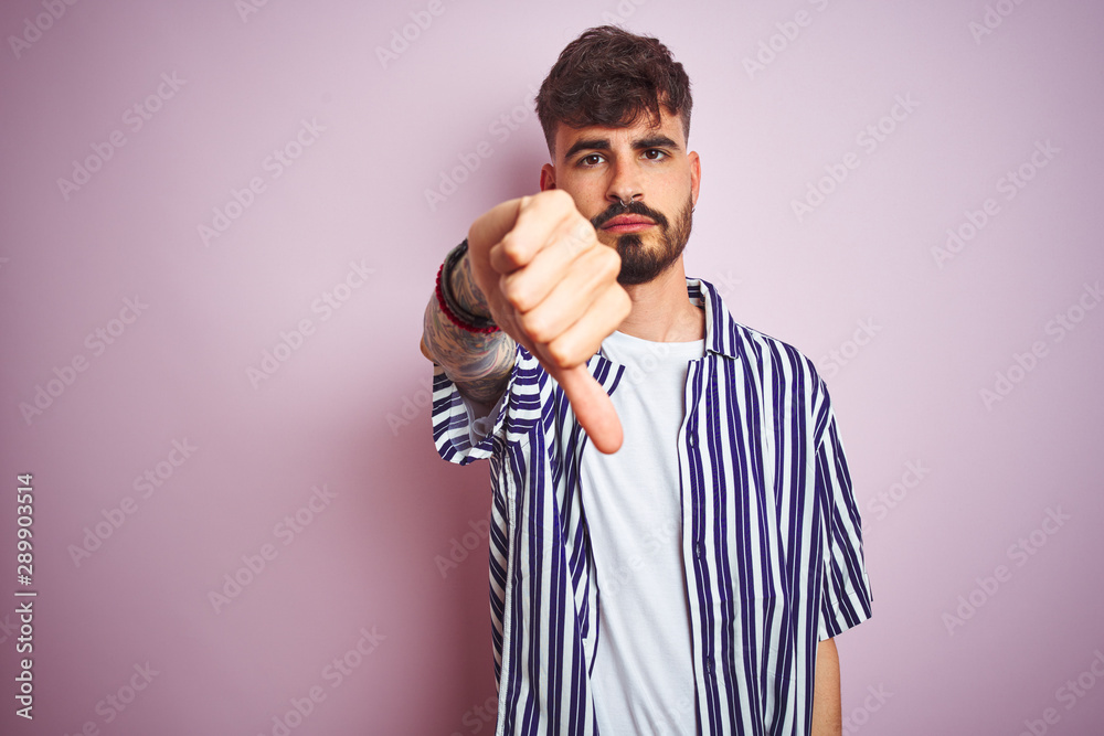 Young man with tattoo wearing striped shirt standing over isolated pink background looking unhappy and angry showing rejection and negative with thumbs down gesture. Bad expression. Young man with tattoo wearing striped shirt standing over isolated pink background looking unhappy and angry showing rejection and negative with thumbs down gesture. Bad expression.