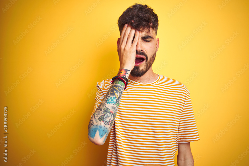 Young man with tattoo wearing striped t-shirt standing over isolated yellow background Yawning tired covering half face, eye and mouth with hand. Face hurts in pain. Young man with tattoo wearing striped t-shirt standing over isolated yellow background Yawning tired covering half face, eye and mouth with hand. Face hurts in pain.