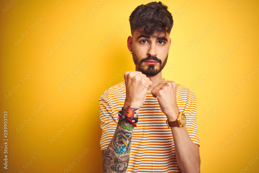 Young man with tattoo wearing striped t-shirt standing over isolated yellow background Ready to fight with fist defense gesture, angry and upset face, afraid of problem Young man with tattoo wearing striped t-shirt standing over isolated yellow background Ready to fight with fist defense gesture, angry and upset face, afraid of problem