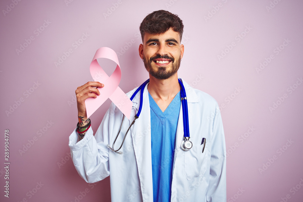 Young doctor man with tattoo holding cancer ribbon standing over isolated pink background with a happy face standing and smiling with a confident smile showing teeth Young doctor man with tattoo holding cancer ribbon standing over isolated pink background with a happy face standing and smiling with a confident smile showing teeth