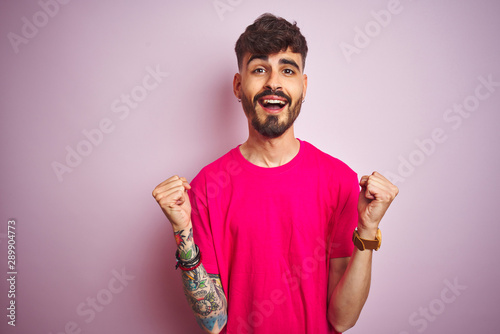 Young man with tattoo wearing t-shirt standing over isolated pink background celebrating surprised and amazed for success with arms raised and open eyes. Winner concept.