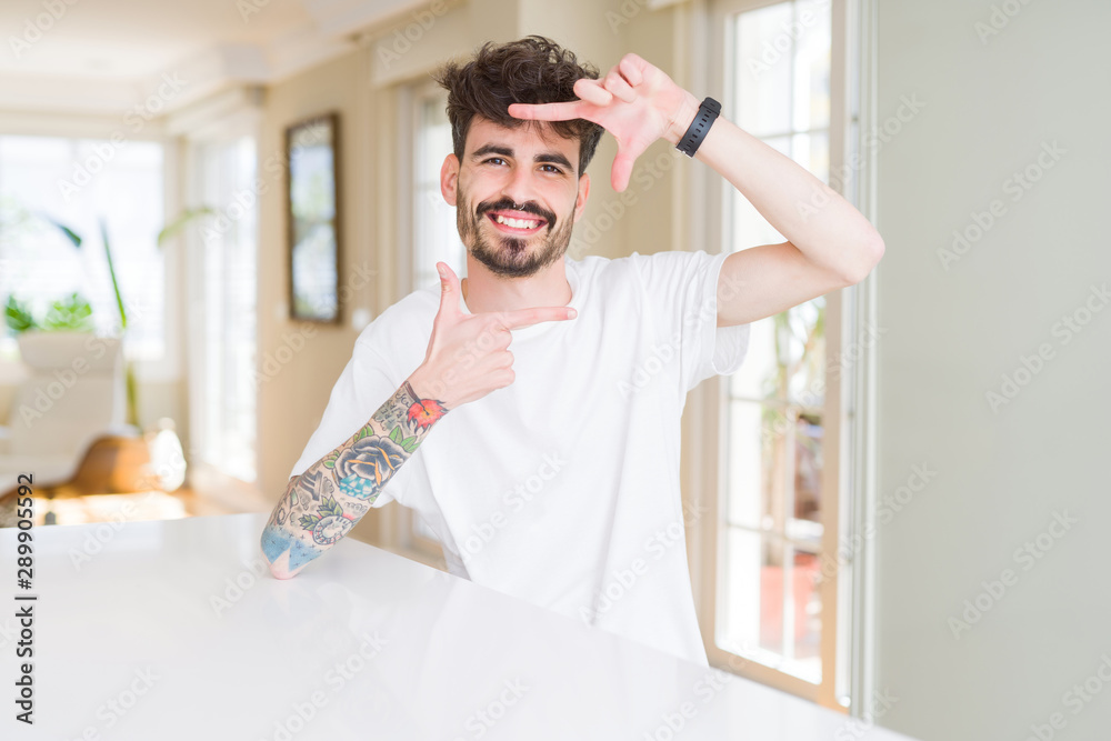 Young man wearing casual t-shirt sitting on white table smiling making frame with hands and fingers with happy face. Creativity and photography concept.