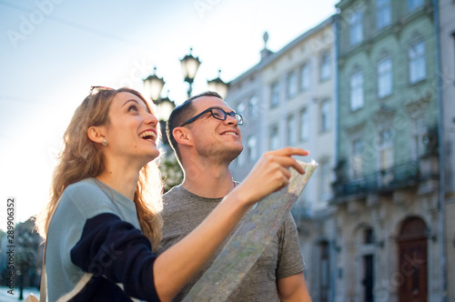 Happy young tourists couple holding a paper map of ancient European city early in the morning on empty square and pointing on some sightseeing place