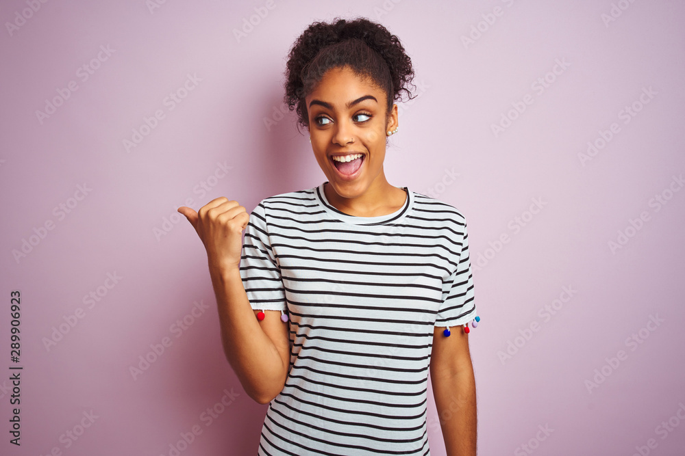 African american woman wearing navy striped t-shirt standing over isolated pink background smiling with happy face looking and pointing to the side with thumb up. African american woman wearing navy striped t-shirt standing over isolated pink background smiling with happy face looking and pointing to the side with thumb up.