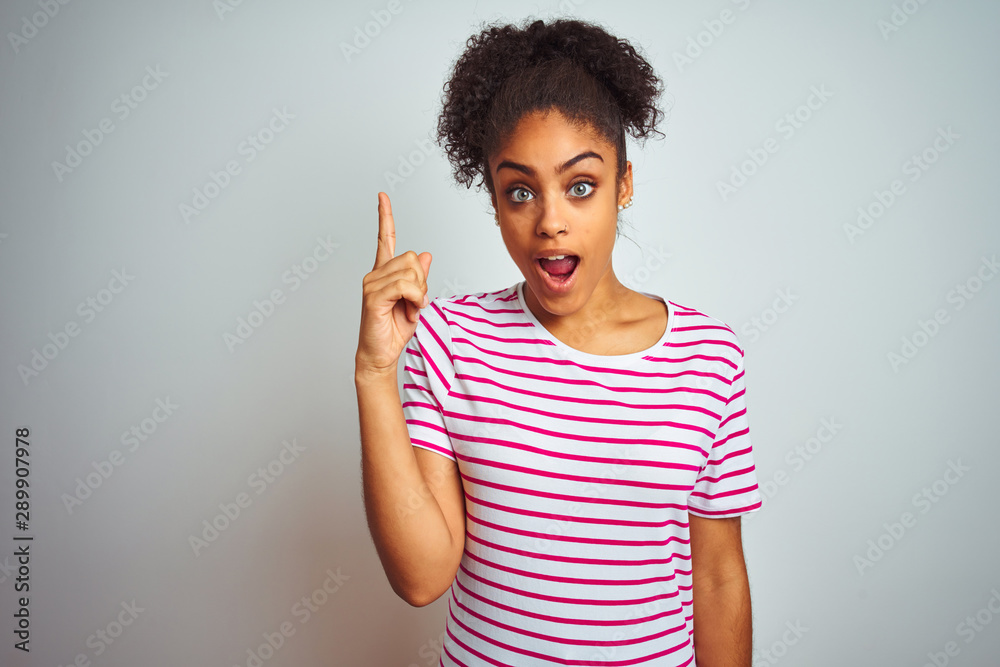 African american woman wearing casual pink striped t-shirt over isolated white background pointing finger up with successful idea. Exited and happy. Number one. African american woman wearing casual pink striped t-shirt over isolated white background pointing finger up with successful idea. Exited and happy. Number one.