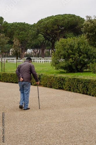 Old man with cane walking towards the white crosses of the soldiers in the  American Cemetery. Sicily – Rome American Cemetery and Memorial in Nettuno Italy.