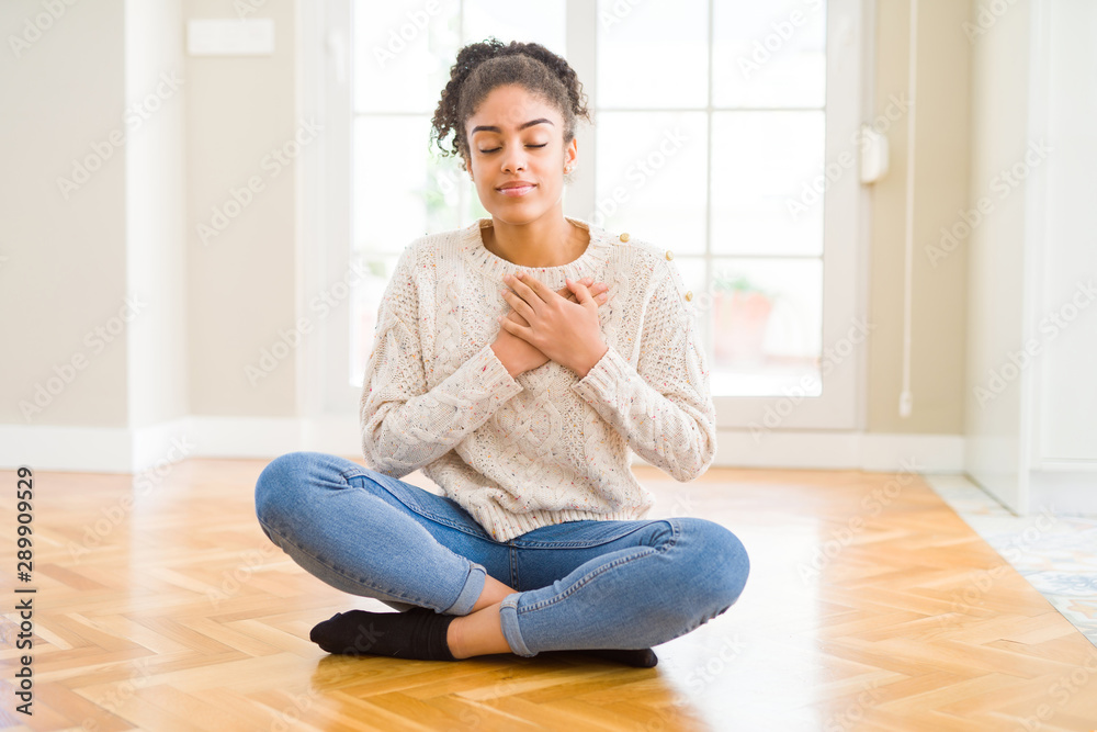 Beautiful young african american woman with afro hair sitting on the floor smiling with hands on chest with closed eyes and grateful gesture on face. Health concept.