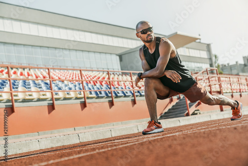 Serious male athlete in sunglasses standing on the running track at the stadium while stretching his legs