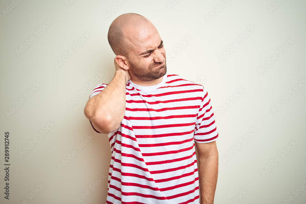 Young bald man with beard wearing casual striped red t-shirt over white isolated background Suffering of neck ache injury, touching neck with hand, muscular pain Young bald man with beard wearing casual striped red t-shirt over white isolated background Suffering of neck ache injury, touching neck with hand, muscular pain