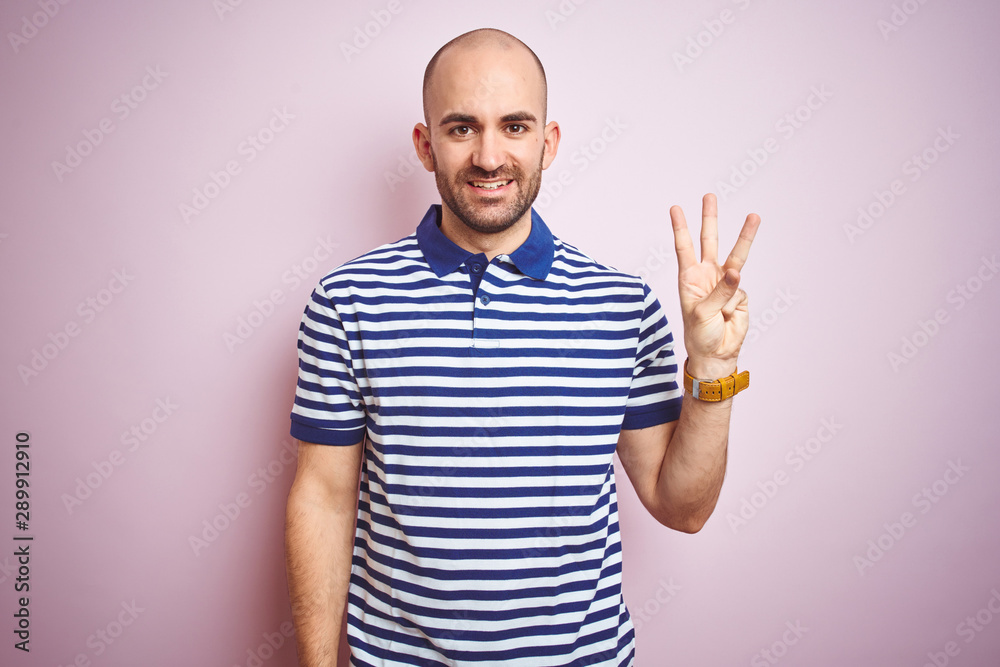 Young bald man with beard wearing casual striped blue t-shirt over pink isolated background showing and pointing up with fingers number three while smiling confident and happy. Young bald man with beard wearing casual striped blue t-shirt over pink isolated background showing and pointing up with fingers number three while smiling confident and happy.