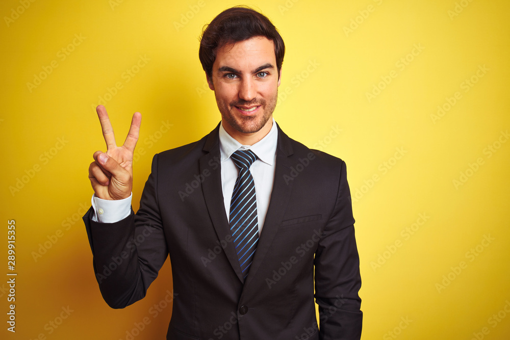 Young handsome businessman wearing suit and tie standing over isolated yellow background showing and pointing up with fingers number two while smiling confident and happy. Young handsome businessman wearing suit and tie standing over isolated yellow background showing and pointing up with fingers number two while smiling confident and happy.