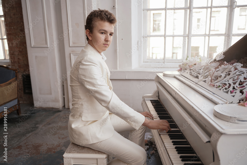 young man teenager in a white business suit with a large white piano in flowers. caucasian male play music at holiday alone.