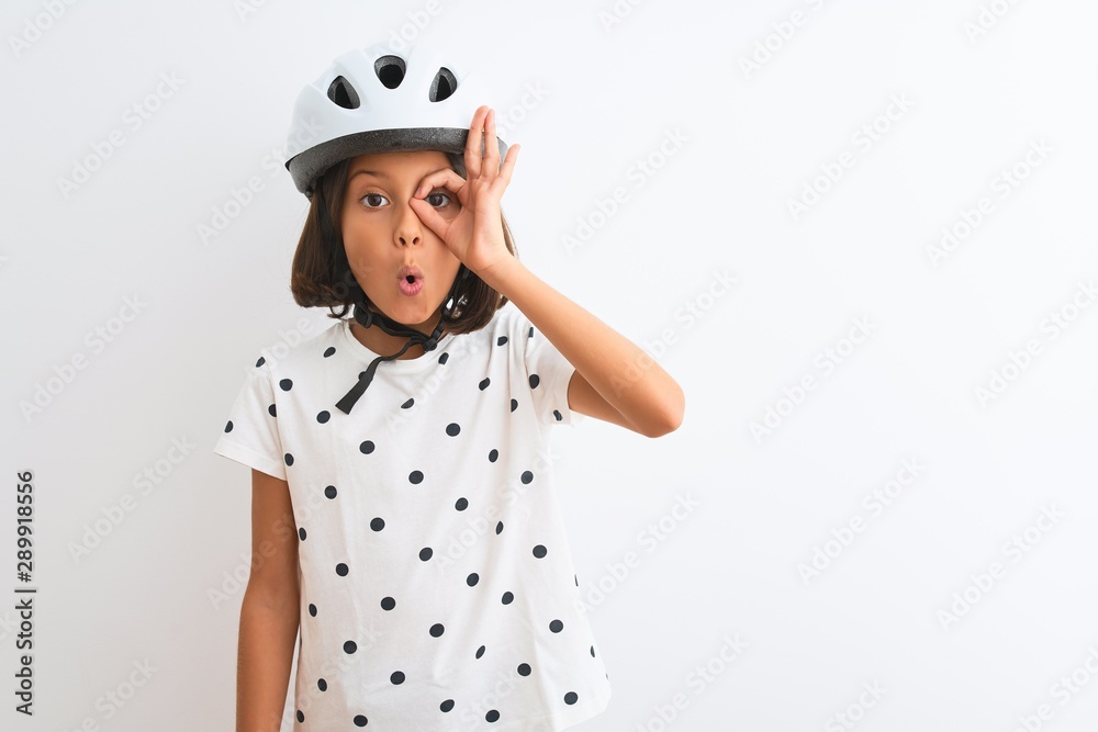 Beautiful child girl wearing security bike helmet standing over isolated white background doing ok gesture shocked with surprised face, eye looking through fingers. Unbelieving expression. Beautiful child girl wearing security bike helmet standing over isolated white background doing ok gesture shocked with surprised face, eye looking through fingers. Unbelieving expression.