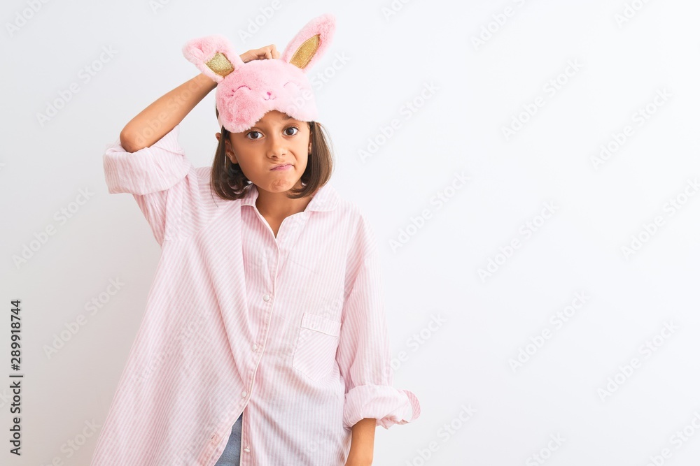 Beautiful child girl wearing sleep mask and pajama standing over isolated white background confuse and wonder about question. Uncertain with doubt, thinking with hand on head. Pensive concept.
