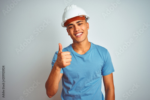 Young brazilian engineer man wearing security helmet standing over isolated white background doing happy thumbs up gesture with hand. Approving expression looking at the camera with showing success.
