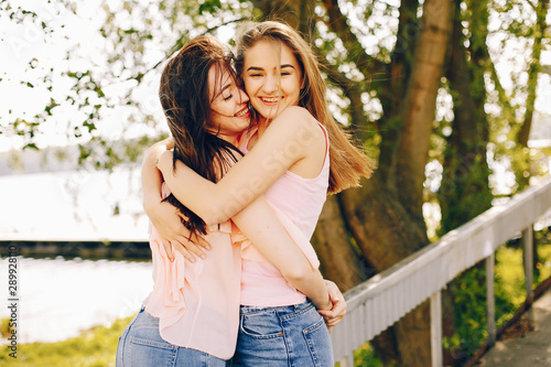 two beautiful and bright friends in pink t-shirts and blue jeans walking in the sunny summer park