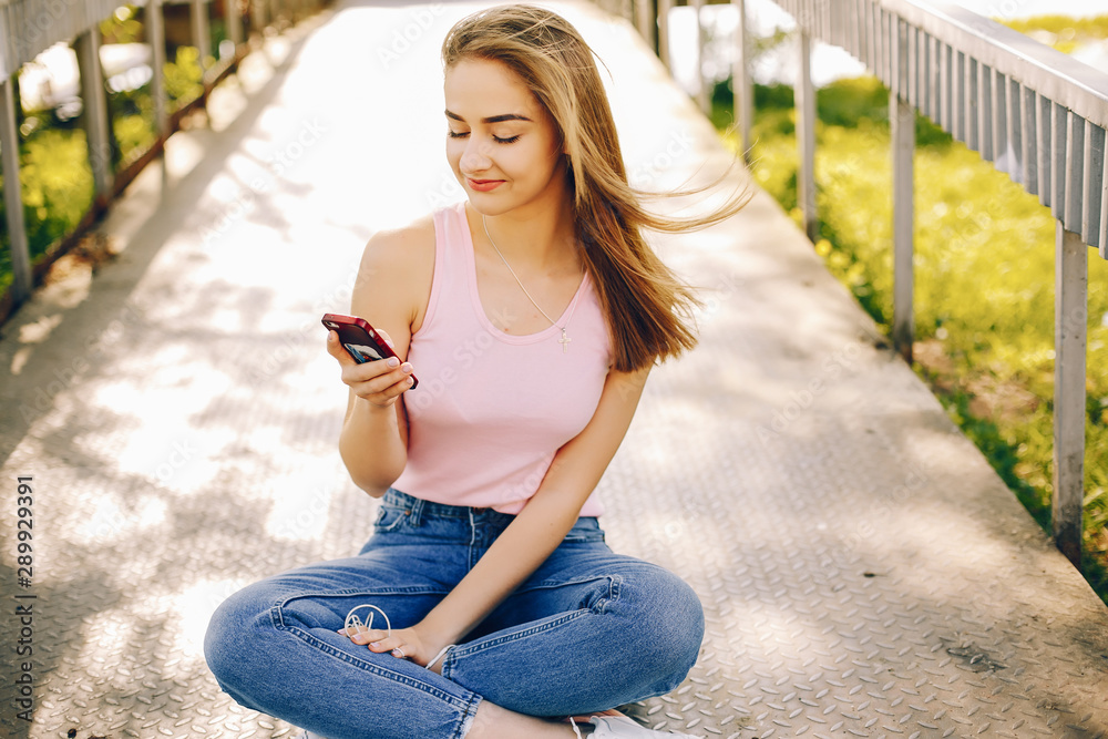beautiful and bright girl in pink t-shirts and blue jeans sitting in the sunny summer park and use the phone with headphones beautiful and bright girl in pink t-shirts and blue jeans sitting in the sunny summer park and use the phone with headphones
