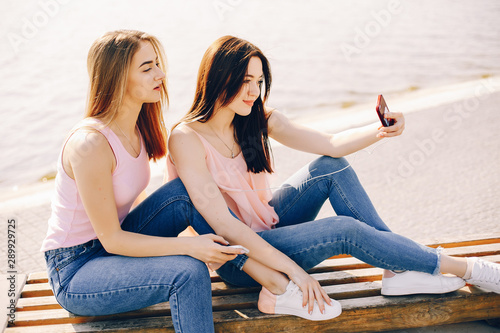 two beautiful and bright friends in pink t-shirts and blue jeans sitting in the sunny summer city and use the phone with headphones © hetmanstock2