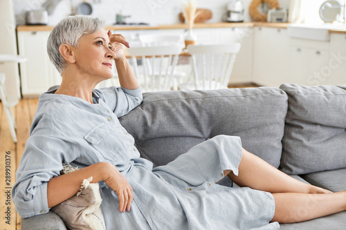 Billede på lærred Side view of attractive stylish European female pensioner in elegant dress sitting barefooted on comfortable couch, having thoughtful look, daydreaming