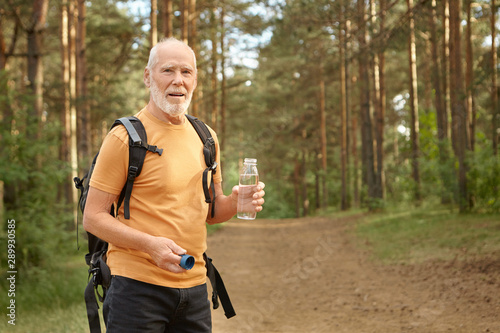 Age, activity, nature and travel. Senior bearded male in hiking outfit standing on path in forest carrying backpack, refreshing himself during trekking, holding bottle of water, looking at camera