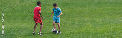 panoramic shot of cute multicultural kids playing football on grass