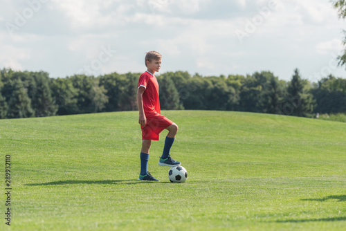 cute kid in sportswear standing on green grass with football