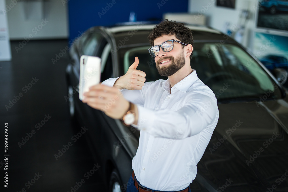 Visiting car dealership. Handsome man is doing selfie with his new car, showing thumb