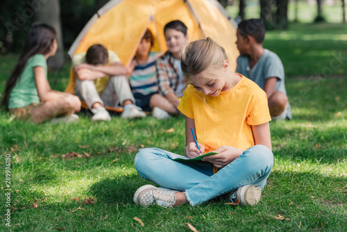 selective focus of happy kid writing in notebook near multicultural friends in camp