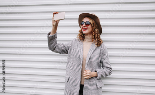Stylish happy smiling woman taking selfie picture by smartphone in gray coat, round hat posing over white wall background © guas