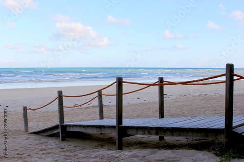 Wooden sidewalk leading to the beach sand. In front the sea and blue sky in the horizon.