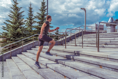 male athlete runs, jogging in morning, doing sports, fitness jogging, workout in city in summer. Free space for text, side view. Steps background, green spruce, blue sky with clouds.