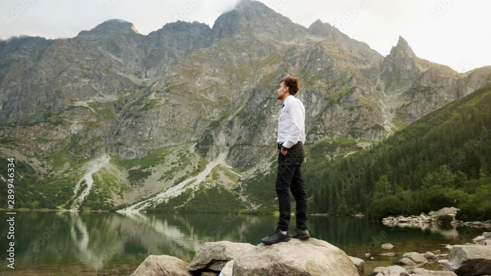 Successful confident businessman standing on the rock near a lake in the mountains at sunset, successful business concept, panorama view