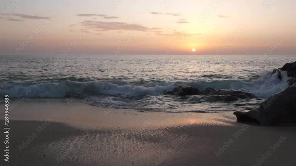 Waves breaking on sandy beach at sunset in slow motion. Sun on horizon in distance and boulders on right. Waves breaking on sandy beach at sunset in slow motion. Sun on horizon in distance and boulders on right.