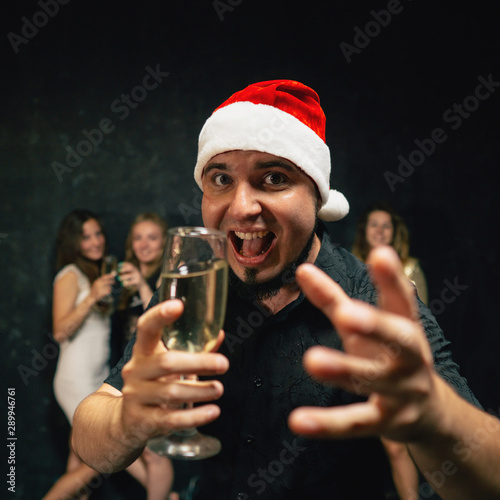 New Year party, holiday, fun. Happy young man in Santa cap drinking champagne invites to join to Christmas celebration