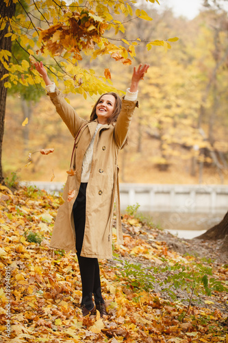 Autumn mood. Romantic woman weared in beige cloak walk in a park on background of beautifull autumn leaves. Smile. fall coming. Brown-haired girl with long hairs. Playing with Yellow leaves in hands