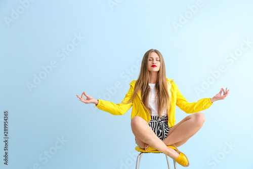 Beautiful young woman meditating on chair against color background