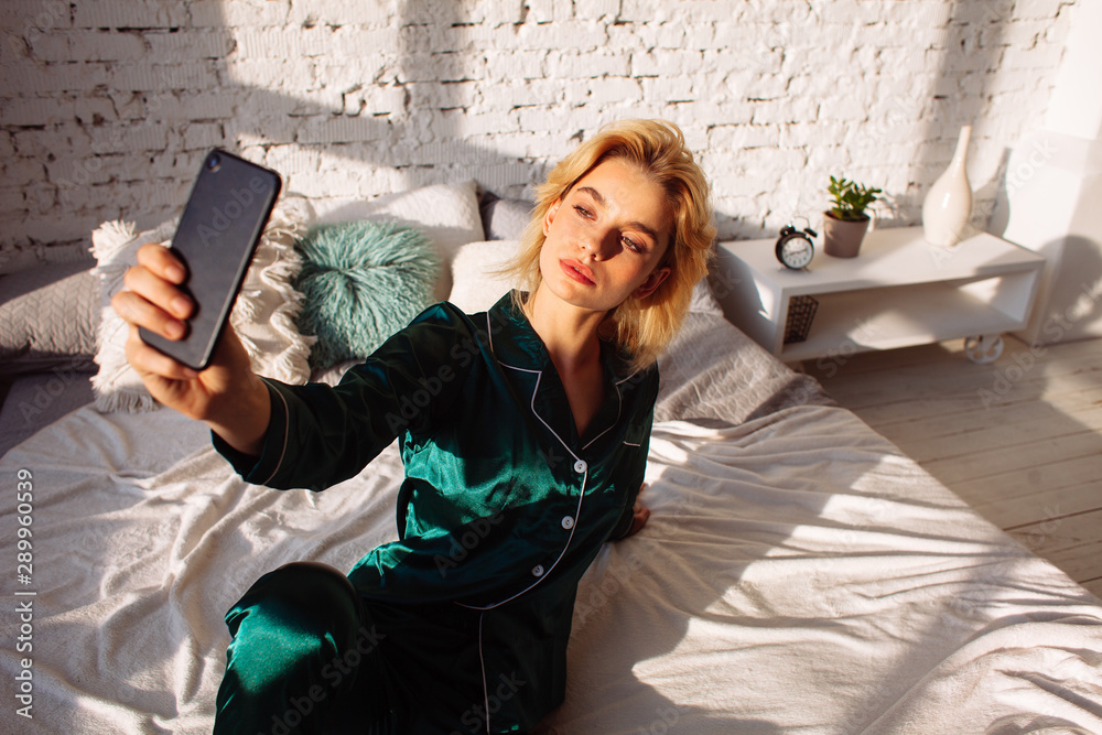 young adult girl in green pajamas taking selfie with smarphone on bed in bedroom interior