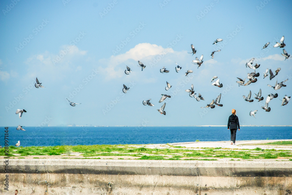 A woman walking under flying pigeons near Aegean sea.