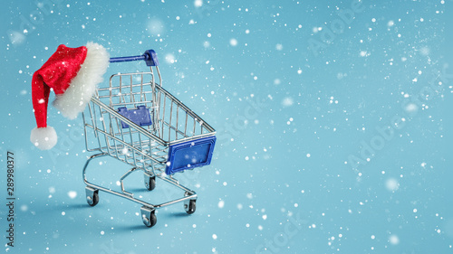 Empty shopping trolley and santa hat on blue snowy background.