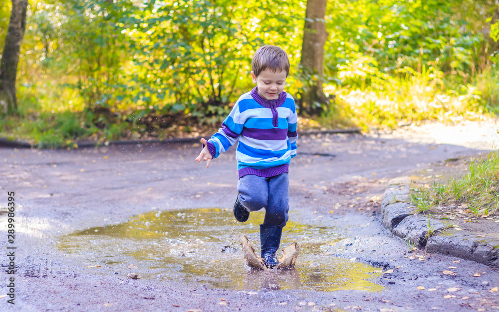 A little boy is jumping in a puddle. A boy in rubber boots. Happy childhood. Puddles after the rain. Warm summer evening. A little boy is jumping in a puddle. A boy in rubber boots. Happy childhood. Puddles after the rain. Warm summer evening.