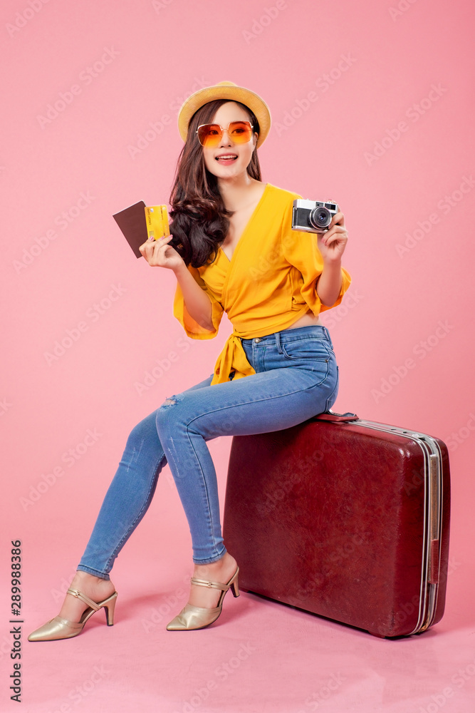 Smiling woman traveler sitting on luggage holding camera passport credit card in holiday on pink backgrounds, relaxation concept, travel concept Smiling woman traveler sitting on luggage holding camera passport credit card in holiday on pink backgrounds, relaxation concept, travel concept